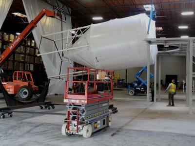 Large white process vessel set on a fabricated steel platform in a warehouse bay, with a scissor lift and a telehandler working beneath it.