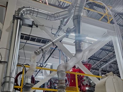Overhead ductwork and process piping run through bar joists on fabricated hangers, branching down toward equipment on the floor below.