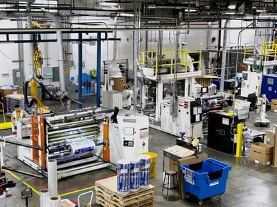 Production line running through an operating plant, with roll stock on pallets, a control console, and yellow-guarded drives along the aisle.