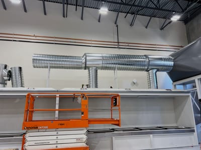 Spiral ductwork branching from a header into the top of a seed dryer body, hung from bar joists above a white powder-coated plenum.