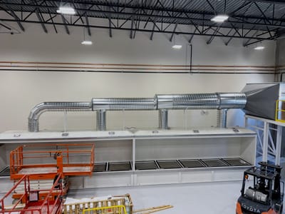 Seed dryer enclosure running the length of a processing bay, with spiral supply ductwork overhead and a scissor lift parked alongside for scale.