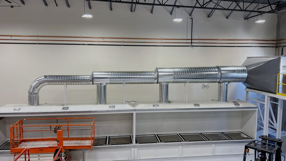 Seed dryer enclosure running the length of a processing bay, with spiral supply ductwork overhead and a scissor lift parked alongside for scale.