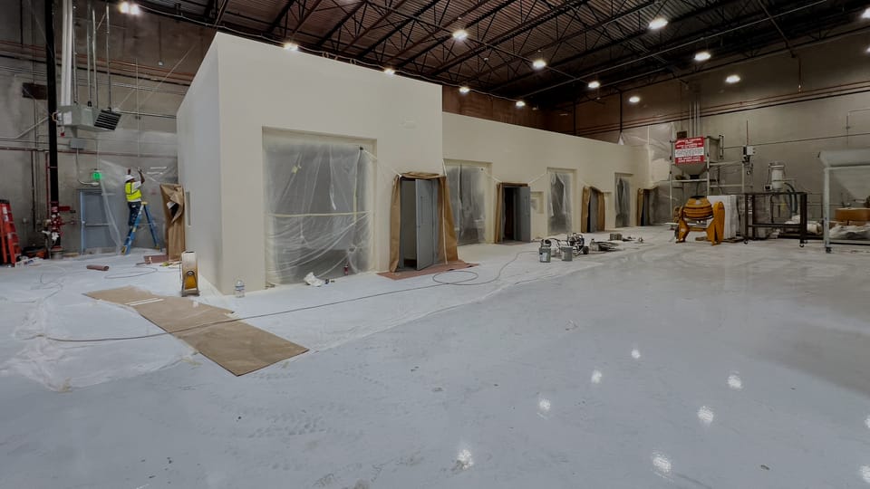 The same enclosure closed in and finished, a smooth-walled room standing free in the warehouse bay with door openings masked for final work.
