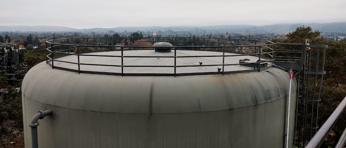 Roof of an in-service industrial water tank with a new perimeter guardrail run around the deck edge, the valley and town below in haze.