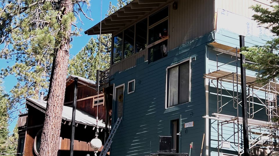 The same house face before the deck, wall sheathing exposed behind scaffold with the crew's truck parked on the access road below.