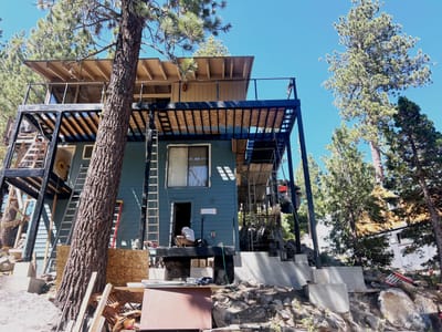 Completed steel deck and balcony wrapping two faces of a hillside house, blackened columns and beams carrying a timber-slatted roof above.