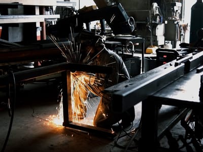 Fabrication bay with steel stock racked overhead and a section being cut on the table, sparks falling to the floor beside the machine.