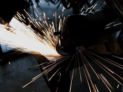Grinder dressing a weld on a steel assembly, a fan of sparks thrown across the frame with the operator's hood and gloves in the near dark.