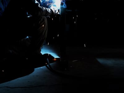 Welder kneeling to run a bead along the base of a fabricated steel frame, hood down and the lead trailing across the shop floor.