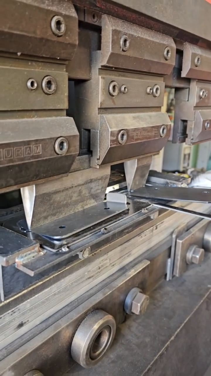 Press brake forming small steel brackets in a spring-loaded welded die, then the formed brackets stacked on a shop bench beside an angle grinder.