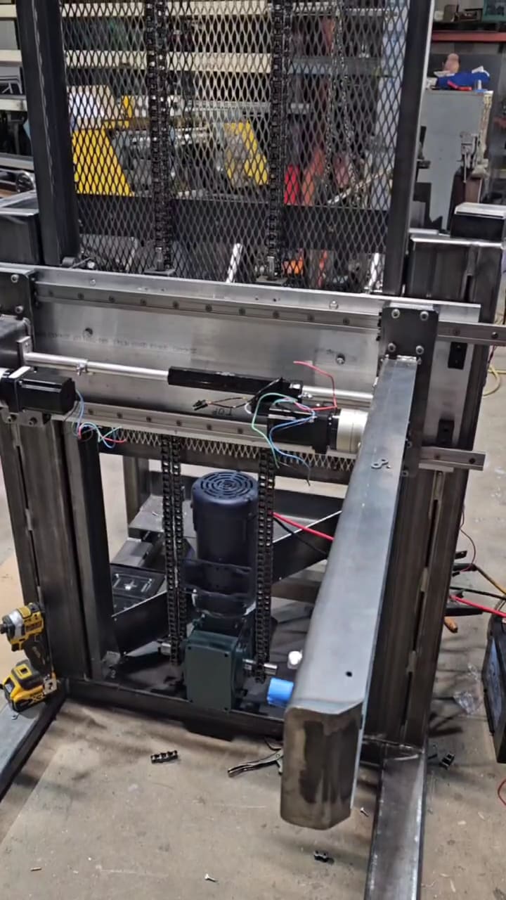 Barrel handler running a lift cycle in the fabrication shop, the carriage travelling up the guarded mast and back down.