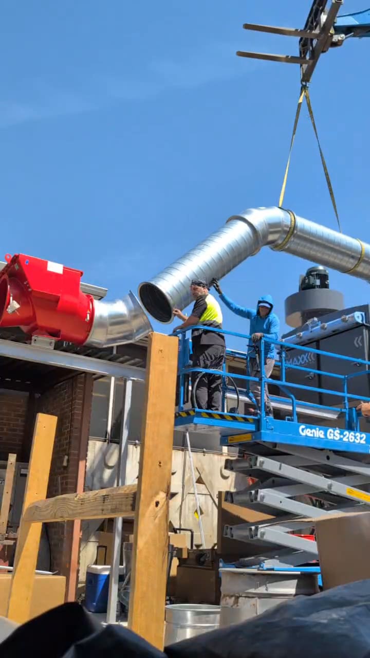 Spiral steel duct elbow suspended on rigging above a scissor lift as a fitter guides it onto a rooftop dust collector.