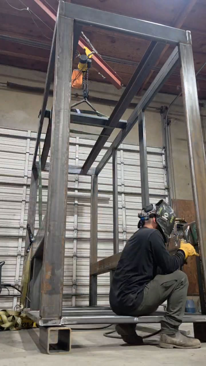 Welder laying a fillet weld at the base of a tall structural steel seed-dryer frame standing in the fabrication bay.