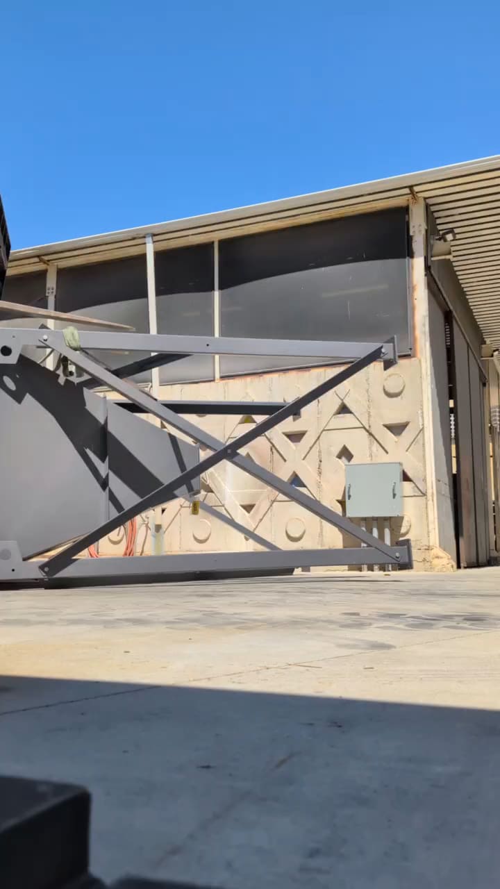 Dark steel hopper on an X-braced leg frame swung upright on overhead rigging outside a shop bay while the crew guides it into place.