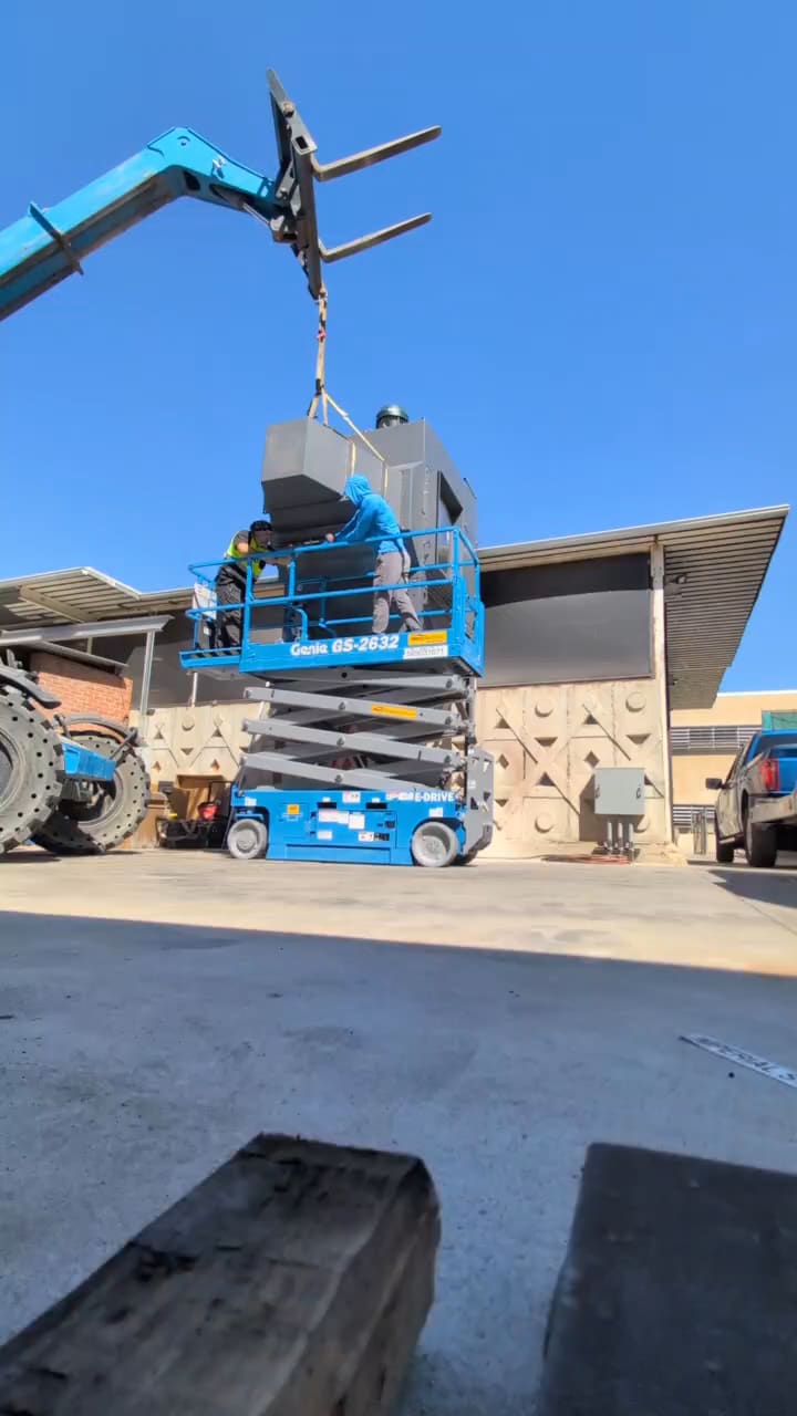 Telehandler lifting a steel equipment section slung on a strap beneath its forks up to two fitters on a scissor lift, against a plant wall.